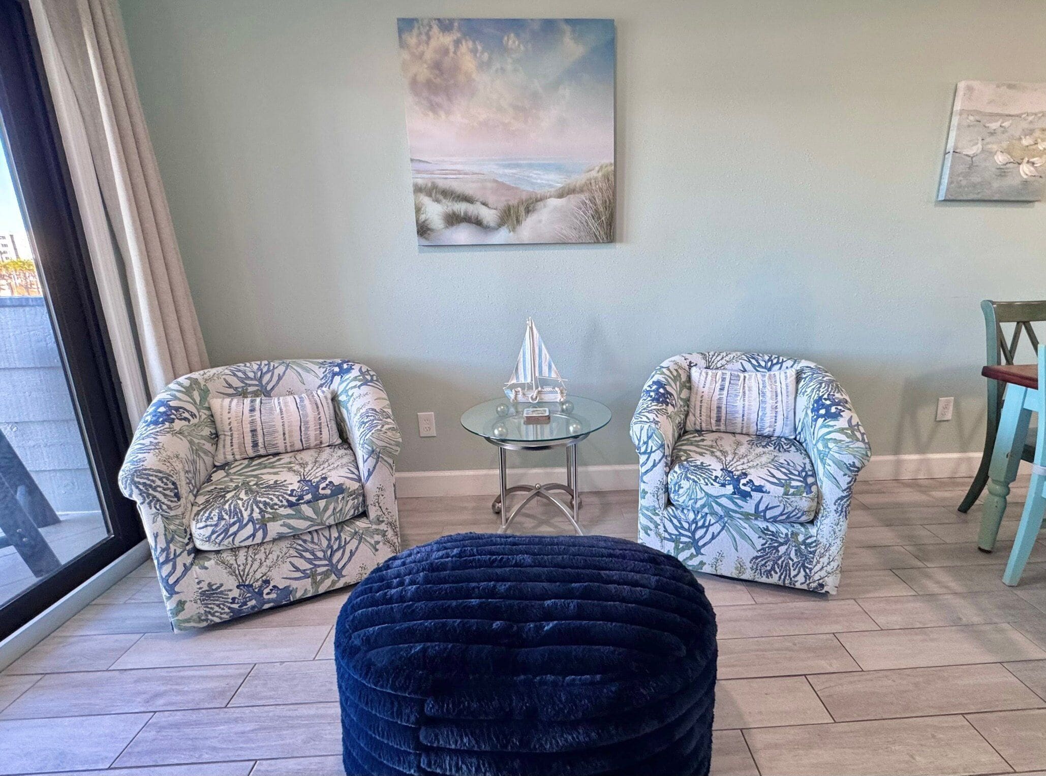 Una sala de estar con dos sillones estampados, una mesa de centro de vidrio y un reposapiés azul.
