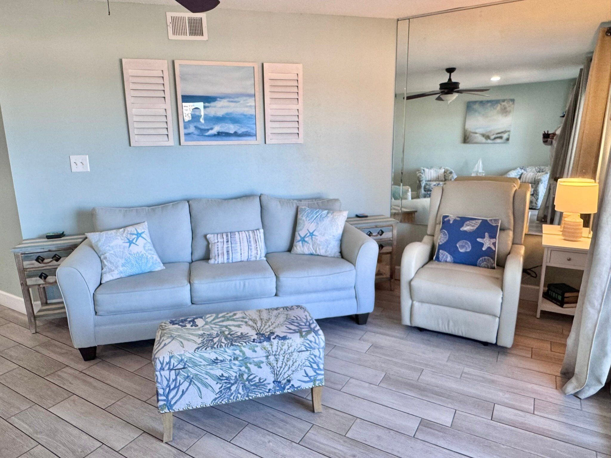 A living room with a light blue sofa, a matching armchair, and a patterned ottoman. There are two wall-mounted artworks depicting ocean scenes.