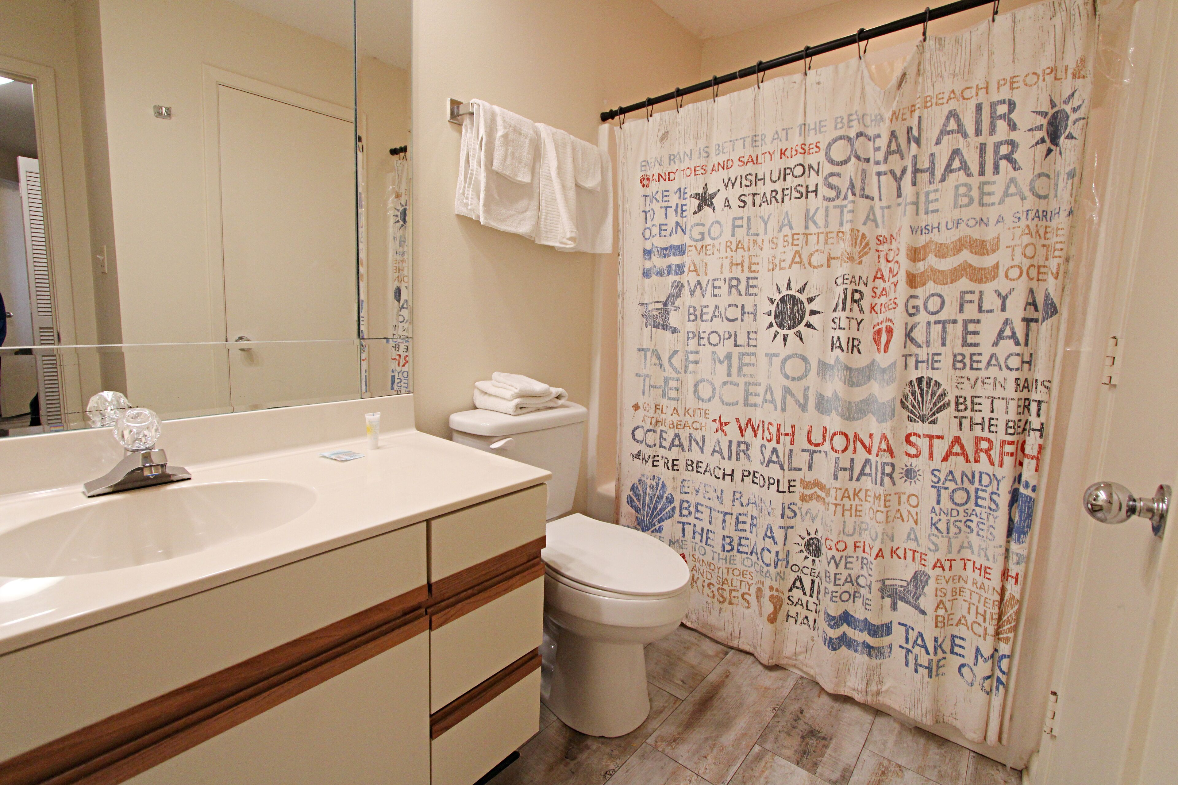 A bathroom with a white sink, mirror, and toilet. There is a shower curtain with text and a towel rack with towels.