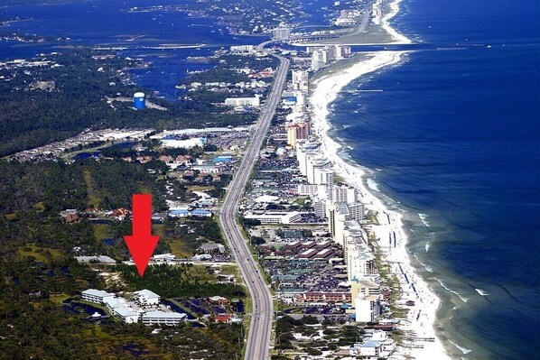 An aerial view of a coastal city with a prominent road running parallel to the beach, a large water tower, and a dense cluster of buildings along the shore.