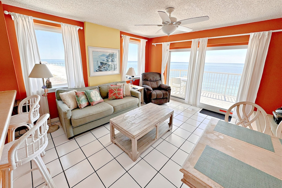 A room with a sofa, armchairs, a table, a ceiling fan, and a view of the sea through large windows.