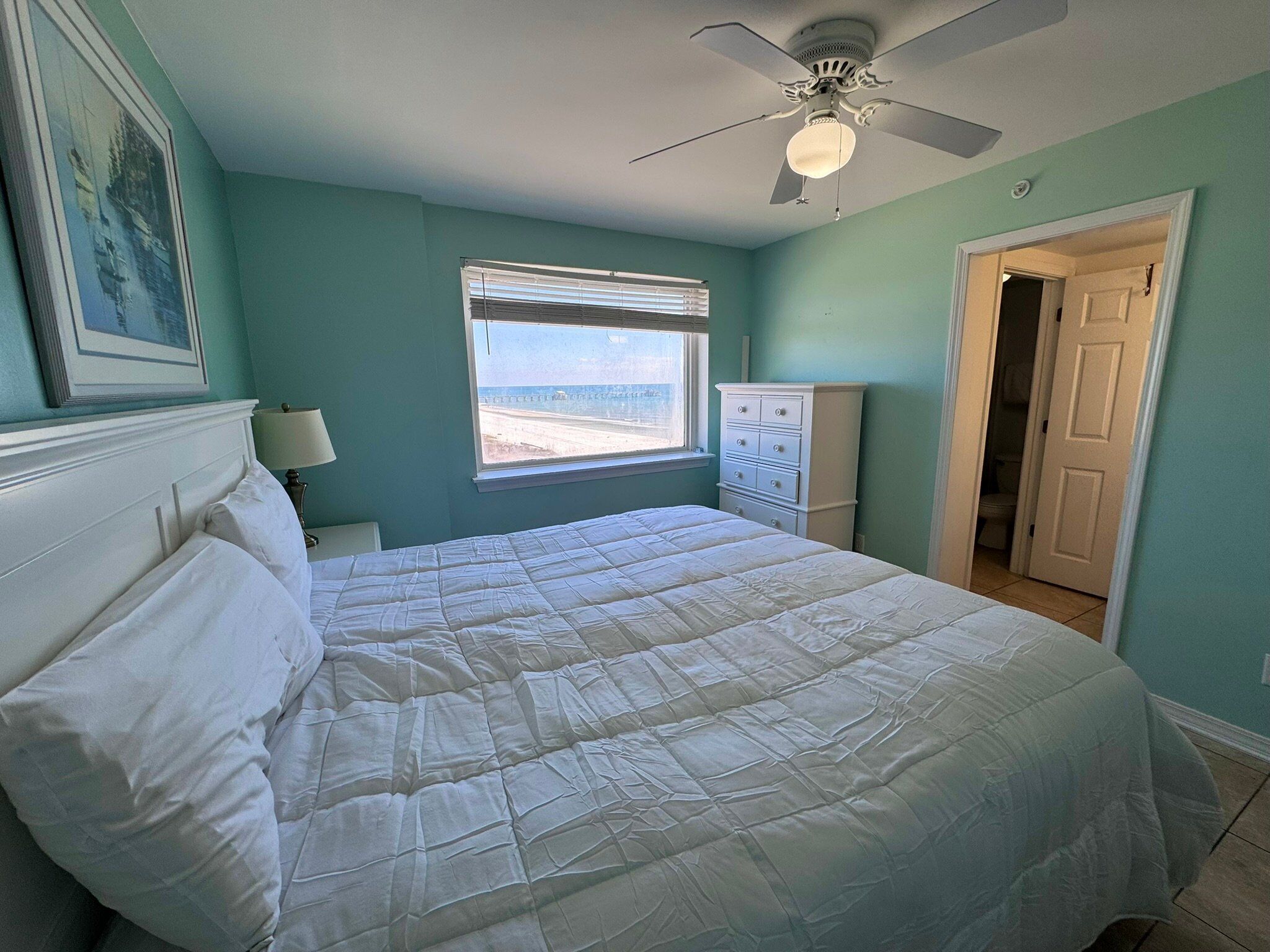 A bedroom with a bed, a ceiling fan, a window with blinds, a dresser, and a door leading to another room.