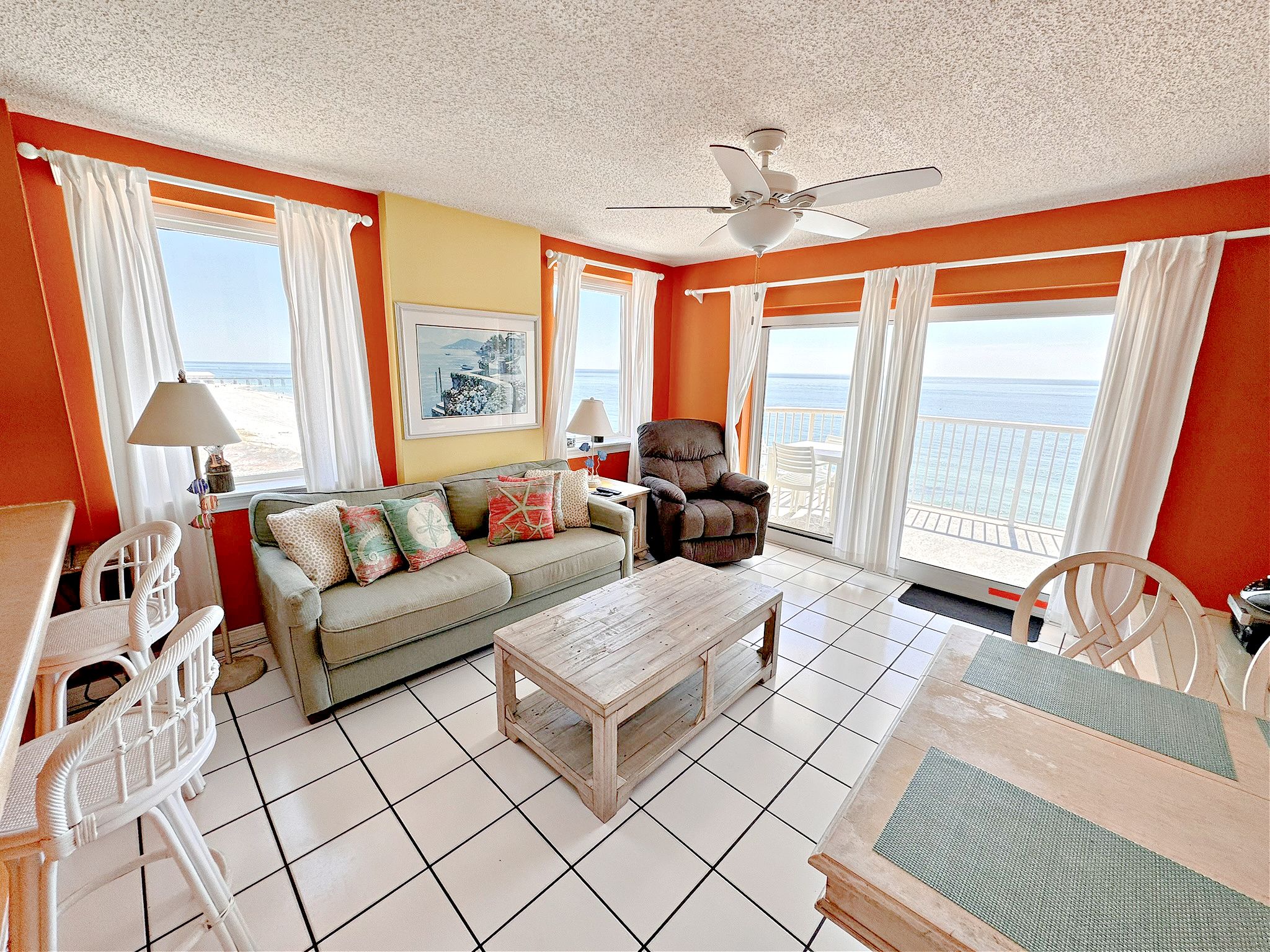 A room with a sofa, armchairs, a table, a ceiling fan, and a view of the sea through large windows.