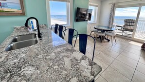 Une cuisine dotée d’un plan de travail en granit, d’un évier et d’un coin repas avec des chaises et une table. Un téléviseur est fixé au mur, et un balcon offre une vue sur l’océan.