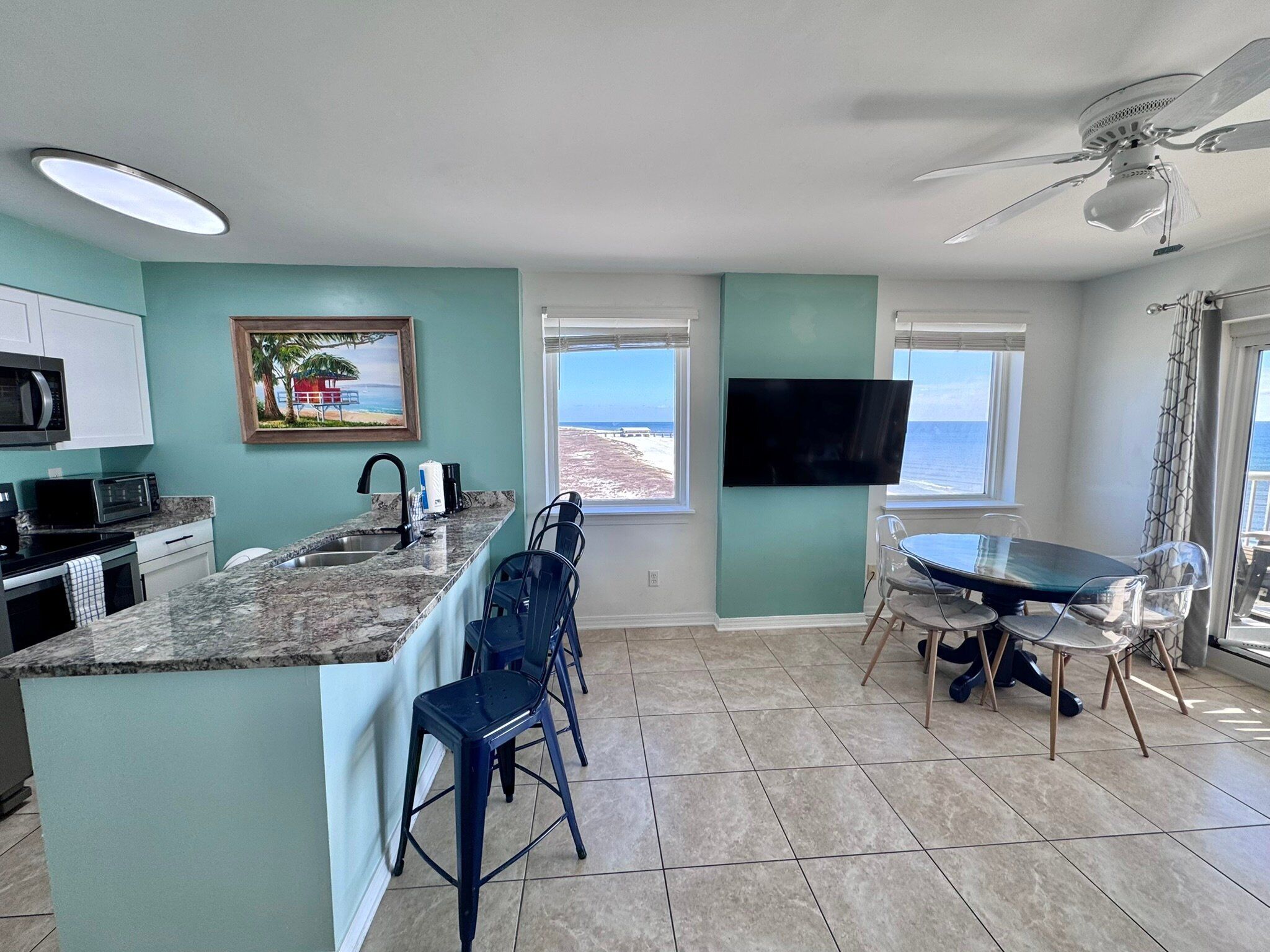 A modern kitchen with a large island, bar stools, a dining table with chairs, a flat-screen TV mounted on the wall, and a view of the ocean through the windows.