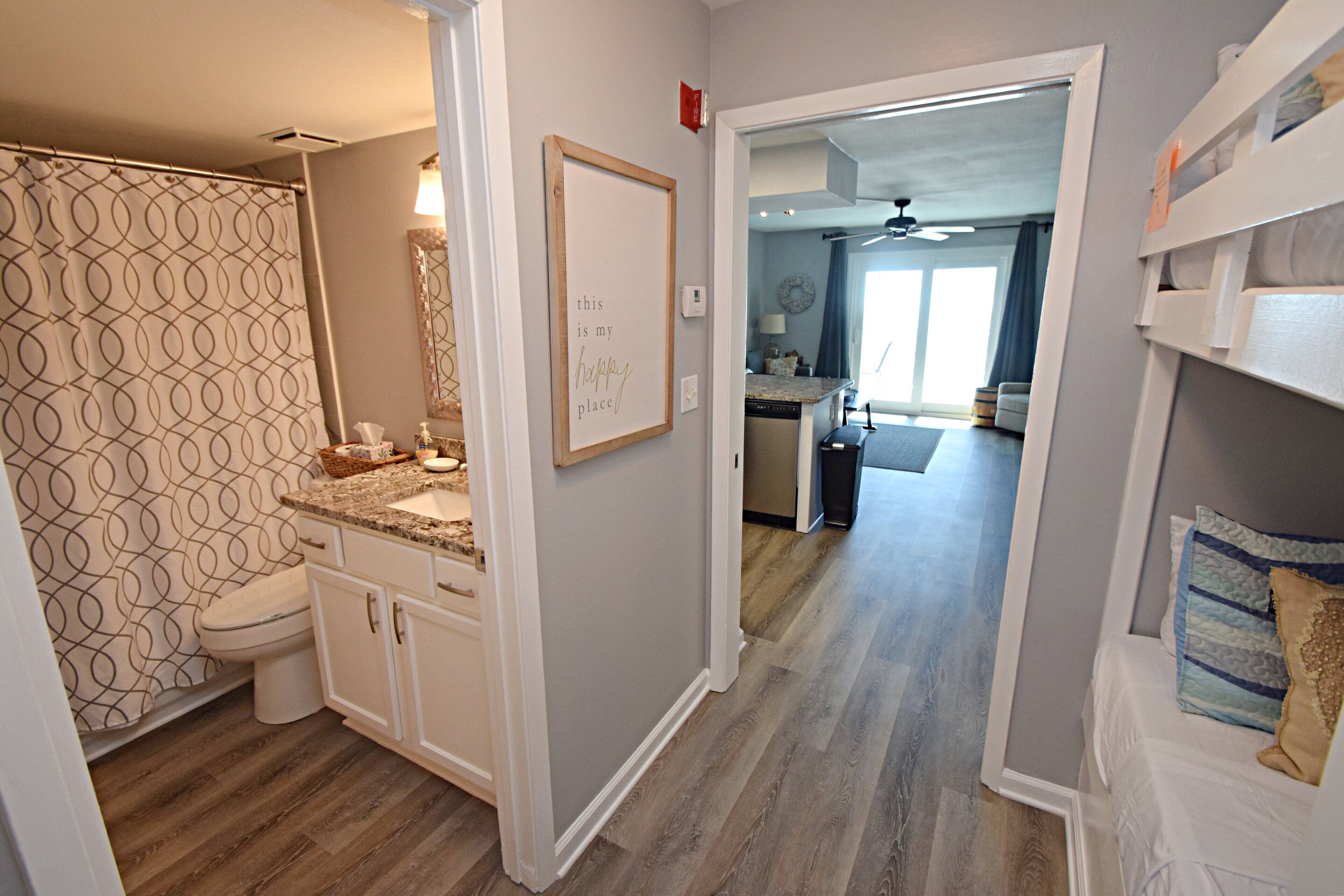 A modern bathroom with a patterned shower curtain, a framed sign on the wall, and a wooden floor.