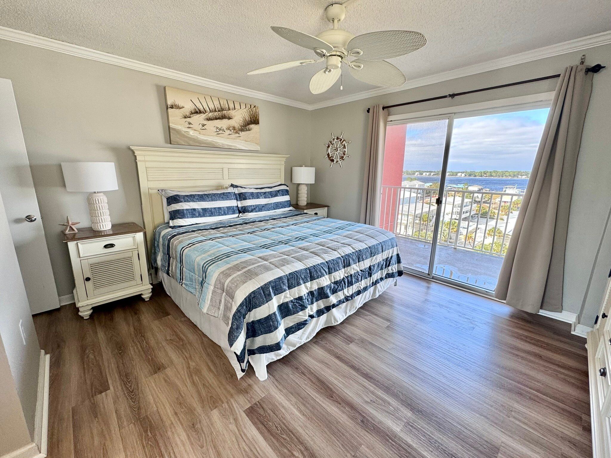 A bedroom with a bed, nightstands, a ceiling fan, a painting, and a balcony with a view.