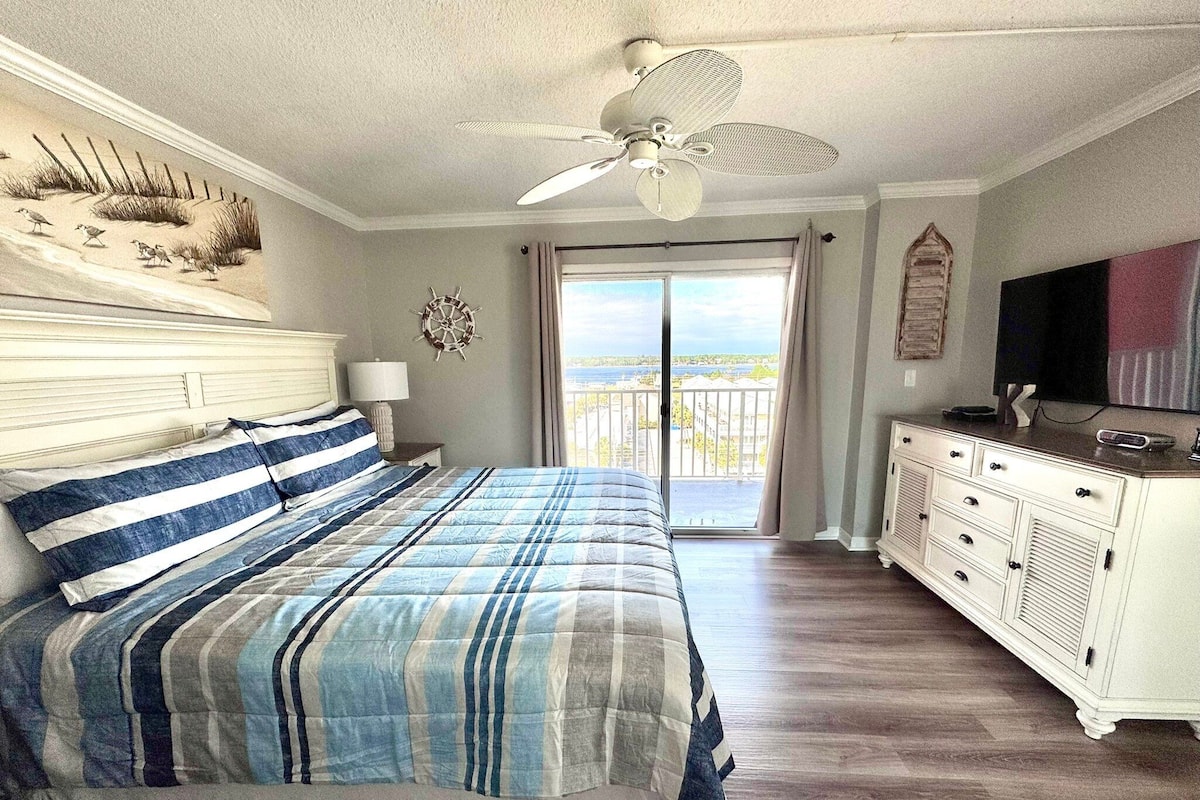 A bedroom with a bed, a dresser, a television, and a view of the outdoors.