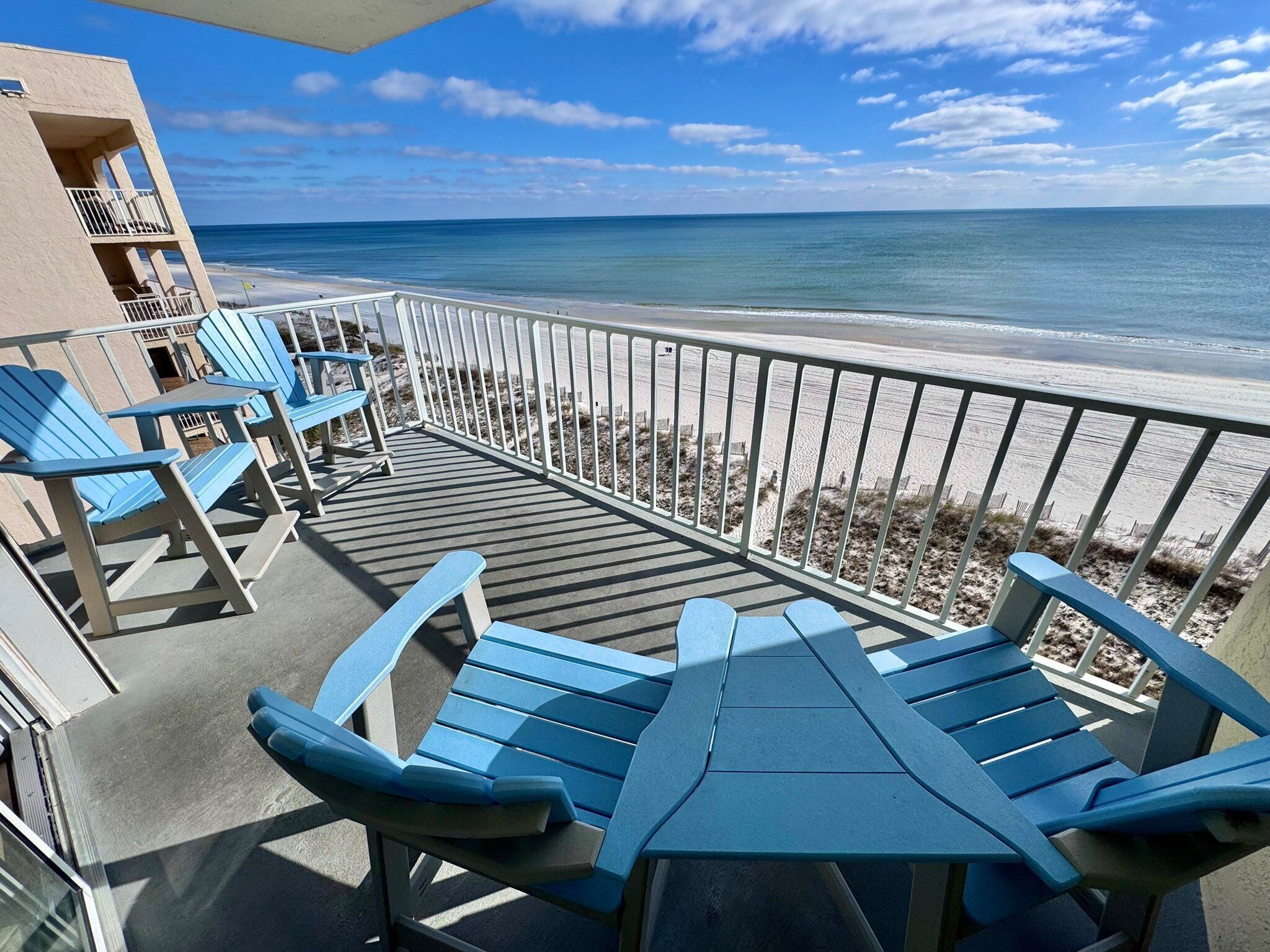 Un balcon avec des fauteuils Adirondack bleus et une table, donnant sur une plage bordée d’une clôture et de dunes de sable.