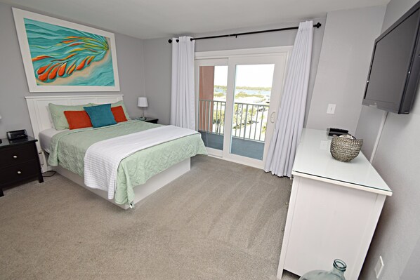 A bedroom with a bed, a desk, a TV, a painting, and a balcony with curtains.