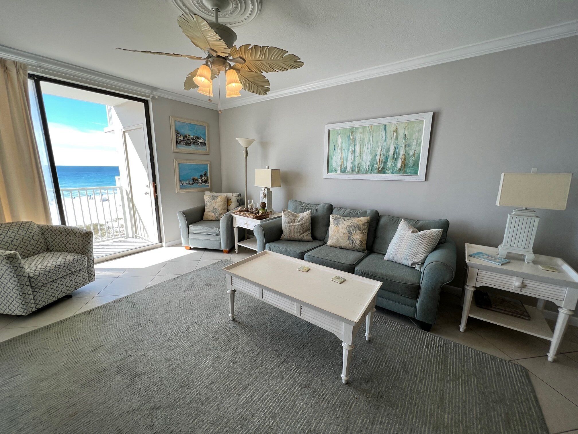 Een woonkamer met een bank, een salontafel, een bijzettafeltje met een lamp en een plafondventilator. Twee schilderijen hangen aan de muur en er is uitzicht op het strand door een schuifdeur.