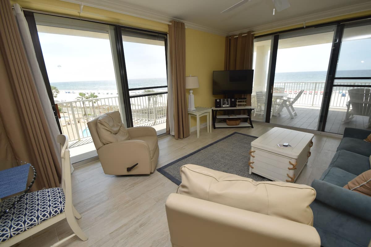 A room with a sofa, armchair, and a TV on a stand, with a view of the sea through large windows.