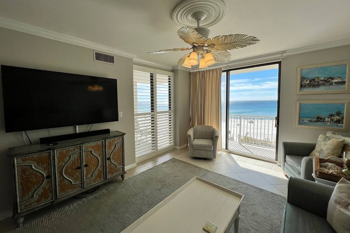A living room with a large flat-screen TV, a ceiling fan, a sofa, and a view of the ocean through large windows.
