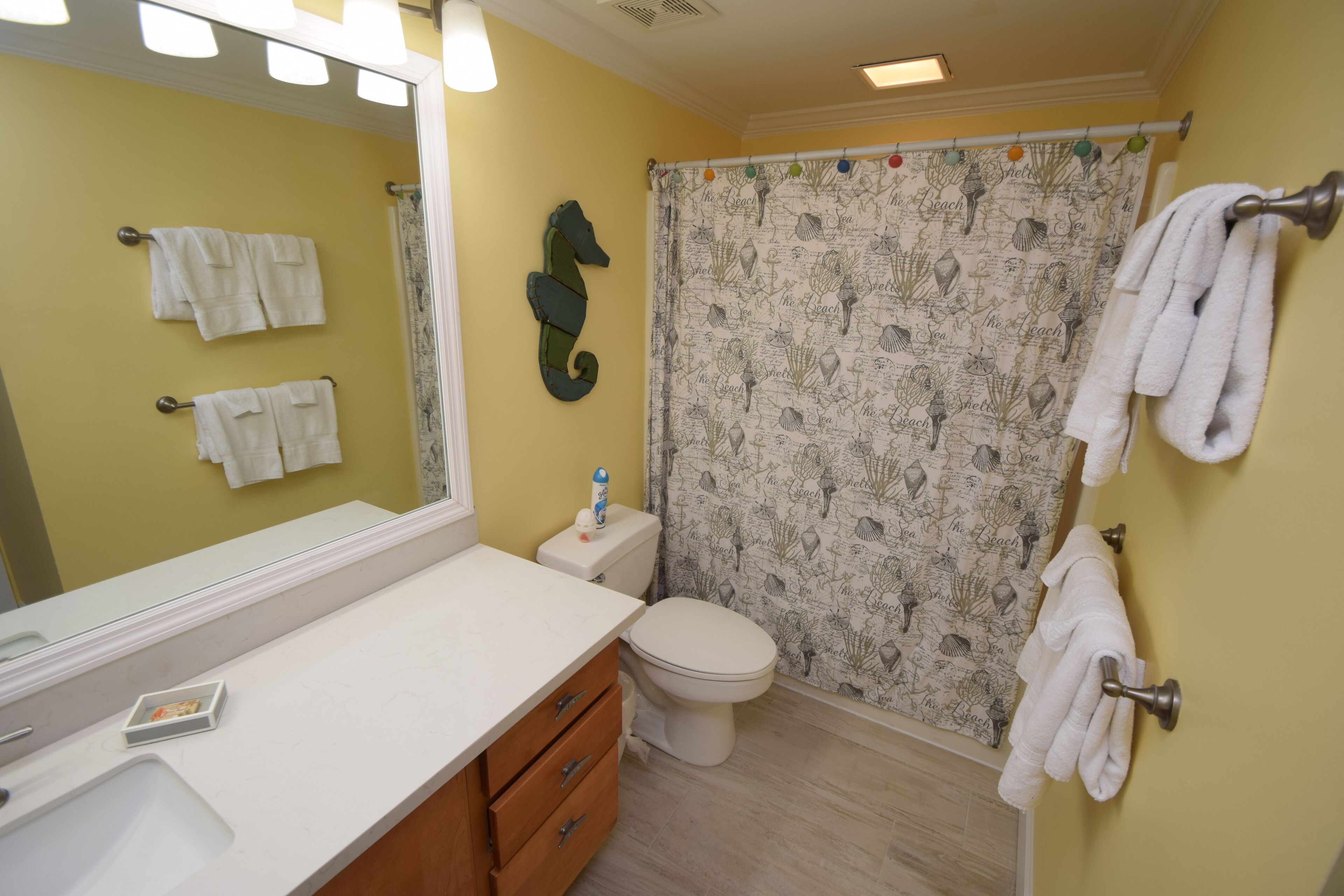 Un baño con lavamanos blanco, tocador de madera, un espejo grande, inodoro y cortina de ducha con motivos náuticos.