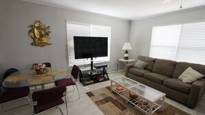 Una sala de estar con una mesa de centro de vidrio, un sofá marrón, un televisor y una mesa de comedor con sillas.