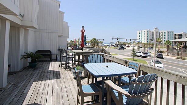 A wooden deck with outdoor seating, a potted plant, and a view of a street with cars and buildings. - Southwinds (Gulf Shores)