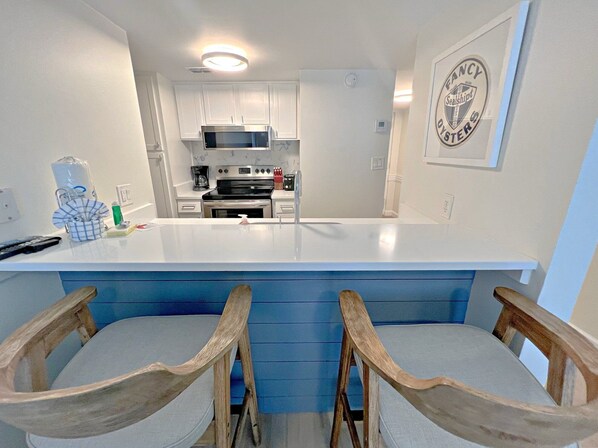 A modern kitchen with white cabinets, a central island, and a microwave. There are two wooden chairs with cushioned seats. - Sugar Beach (Orange Beach)