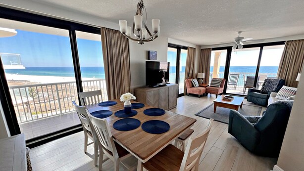 A spacious living area with a balcony overlooking the beach, a dining table with blue placemats, and modern furniture. - Summerhouse (Orange Beach)