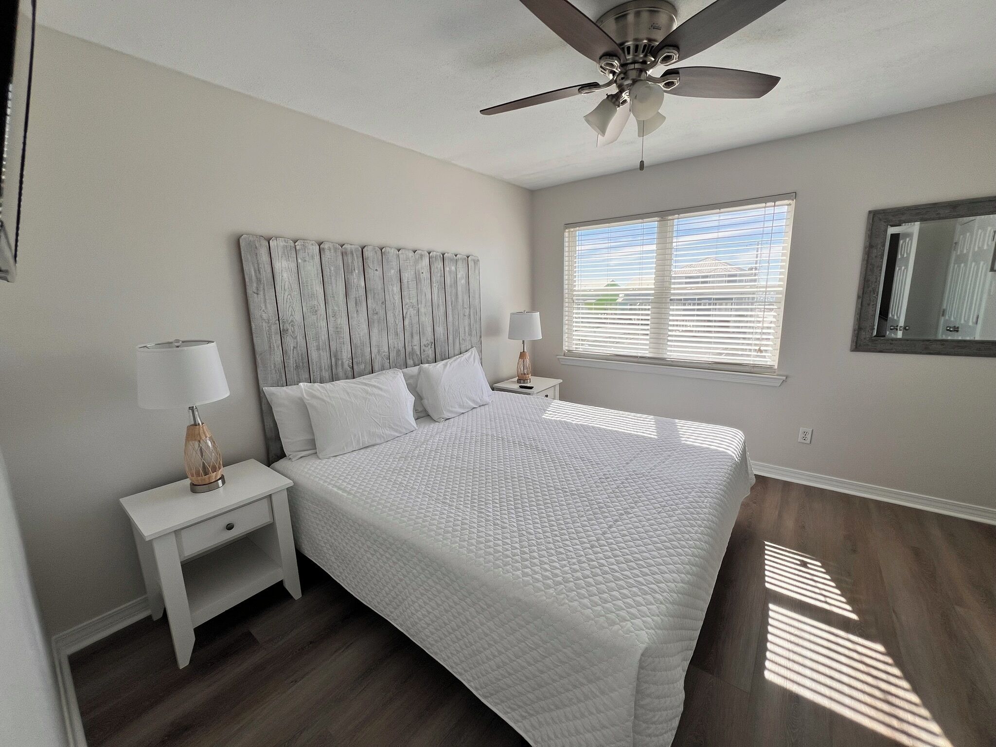 A bedroom with a bed, nightstands, a ceiling fan, a window with blinds, and a mirror on the wall.