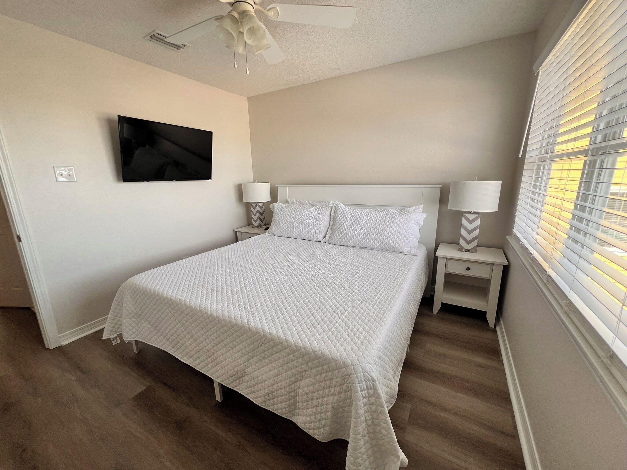 A bedroom with a bed, two bedside tables with lamps, a ceiling fan, a TV mounted on the wall, and a window with blinds.