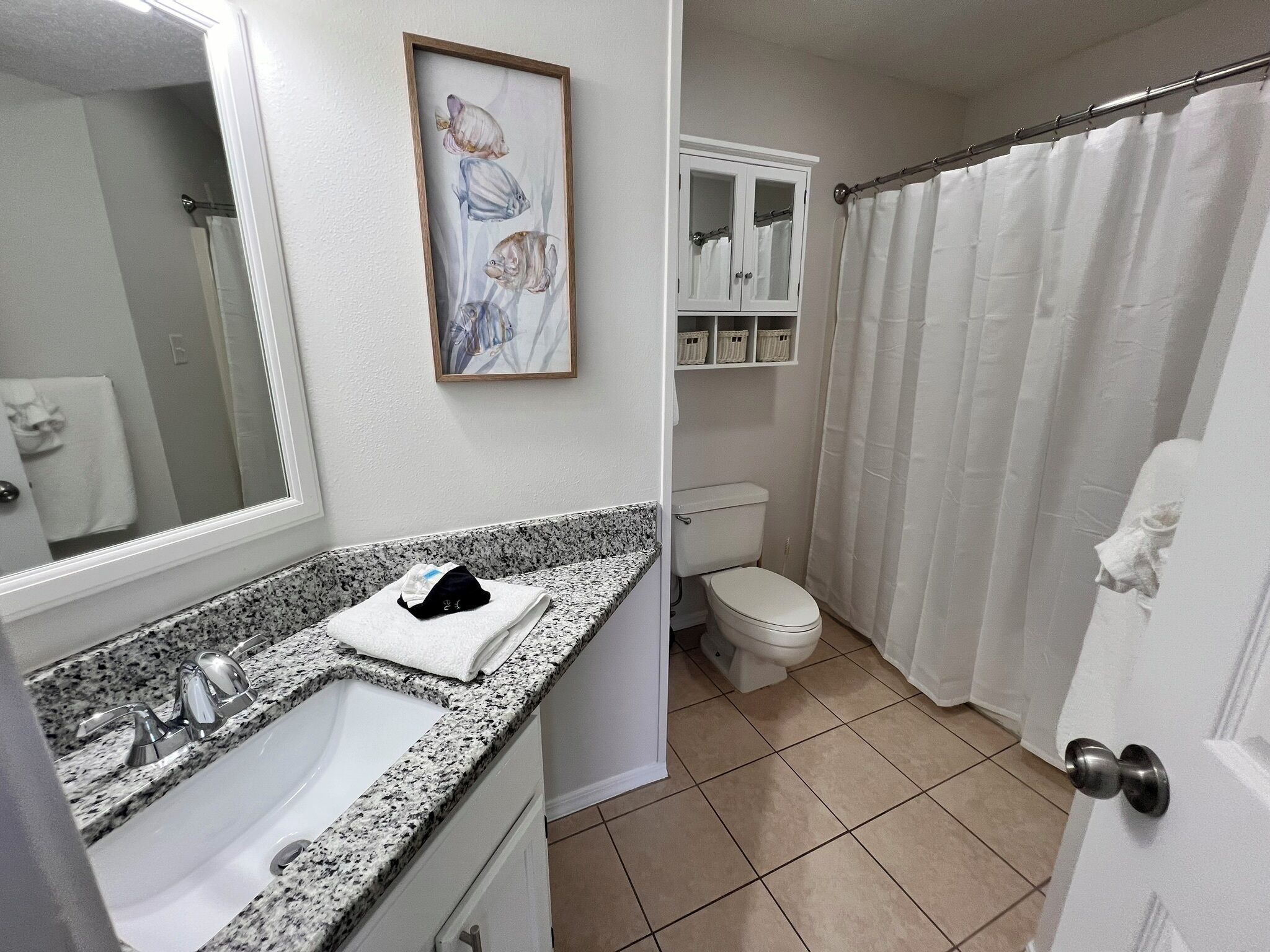 A bathroom with a granite countertop sink, a toilet, a shower curtain, and a framed fish painting on the wall.