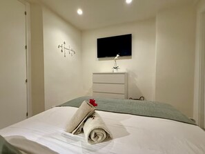 A bedroom with a bed, a flat-screen TV mounted on the wall, a dresser, and a wall-mounted light fixture.