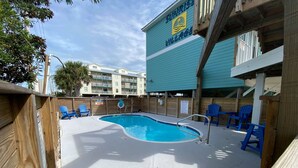 A swimming pool with blue chairs, a wooden deck, and a building with a sign reading "Sunrise Village". - Sunrise Village (Gulf Shores)