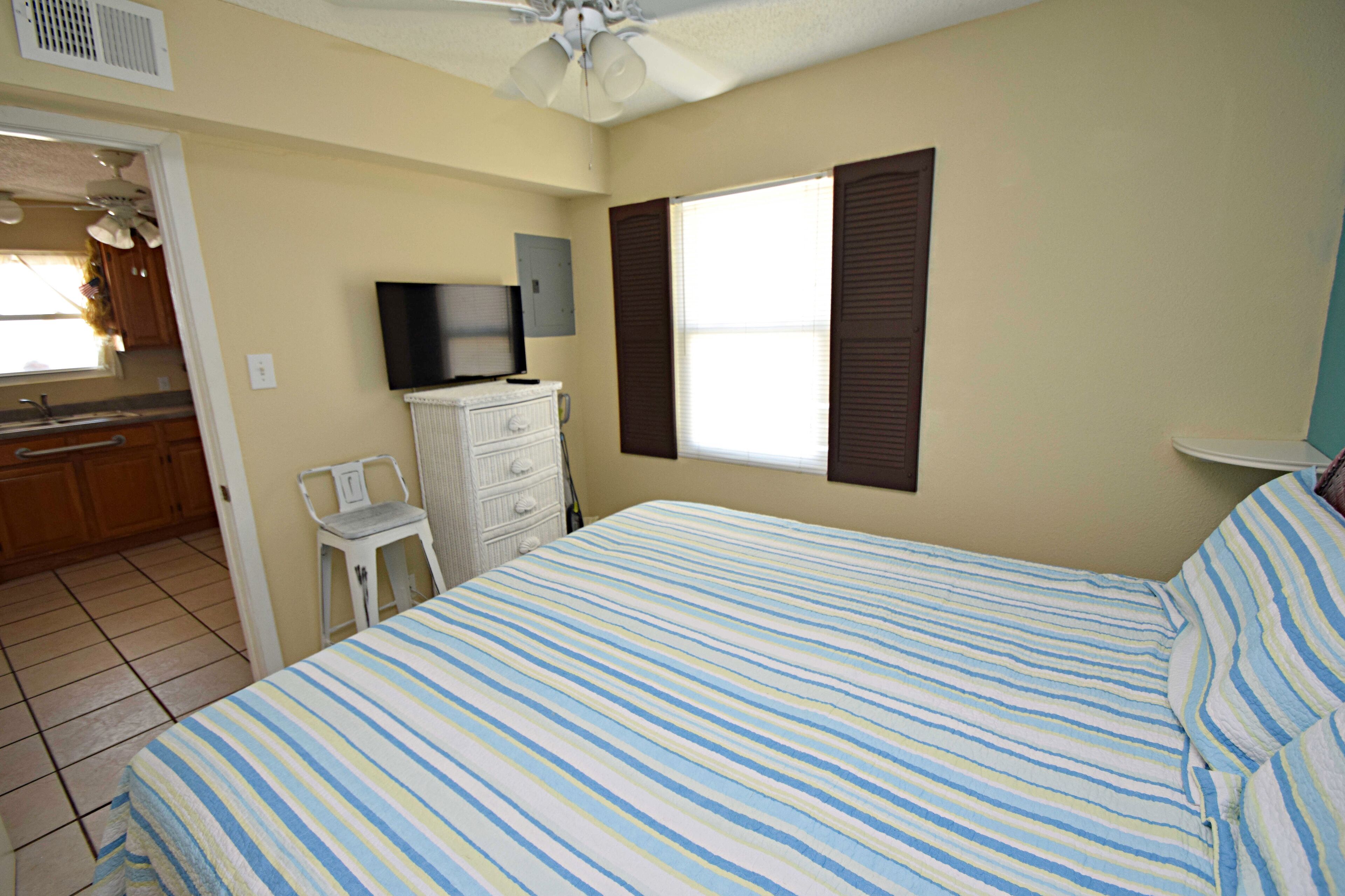 A bedroom with a bed, a television, a window with shutters, a chair, and a kitchenette in the background.