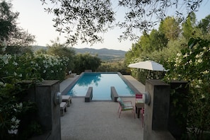Piscina retangular com vista para colinas distantes, cercada por vegetação e estruturas de pedra.