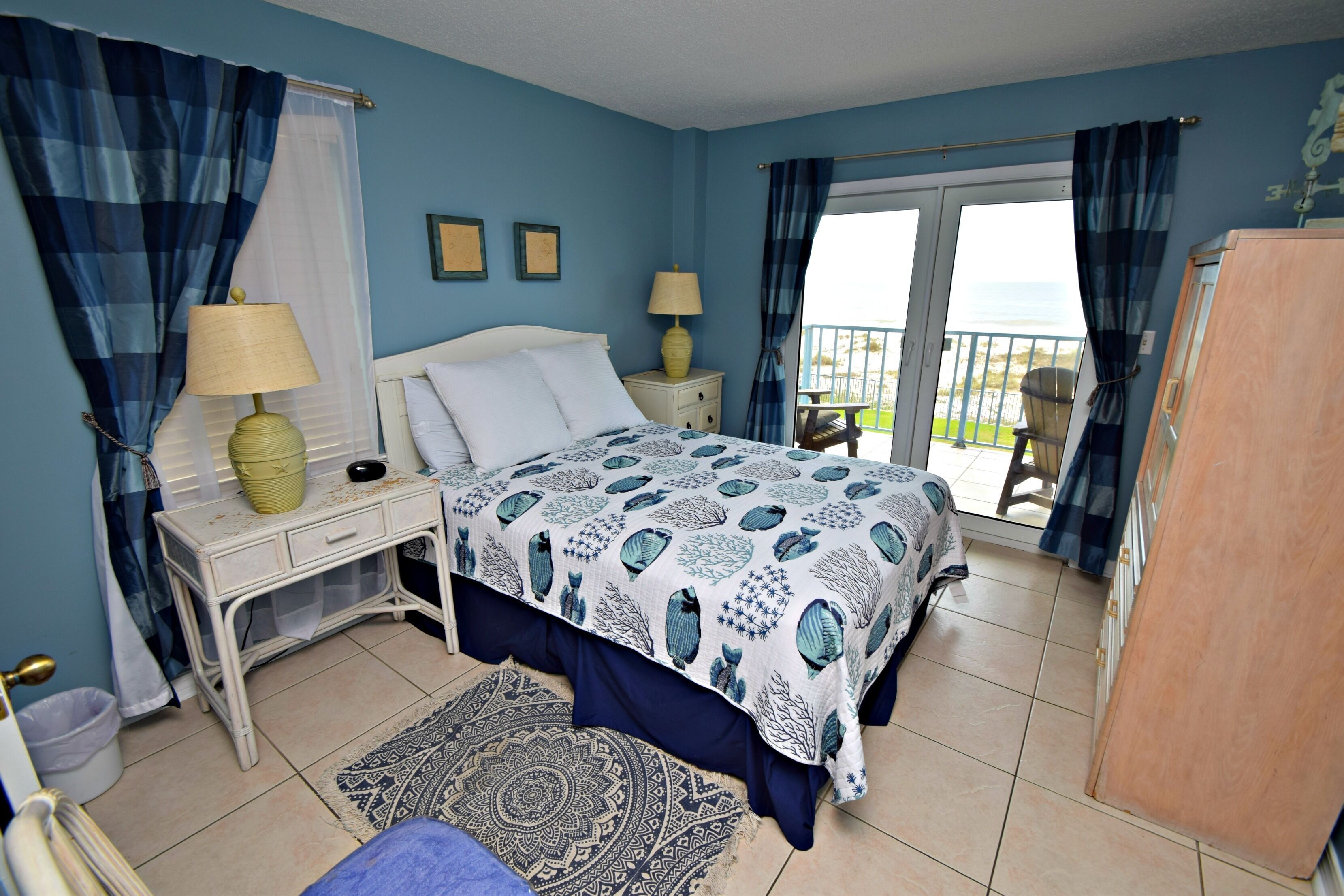 A bedroom with a bed, two bedside tables, a nightstand, a dresser, a patterned rug, and a balcony with curtains.