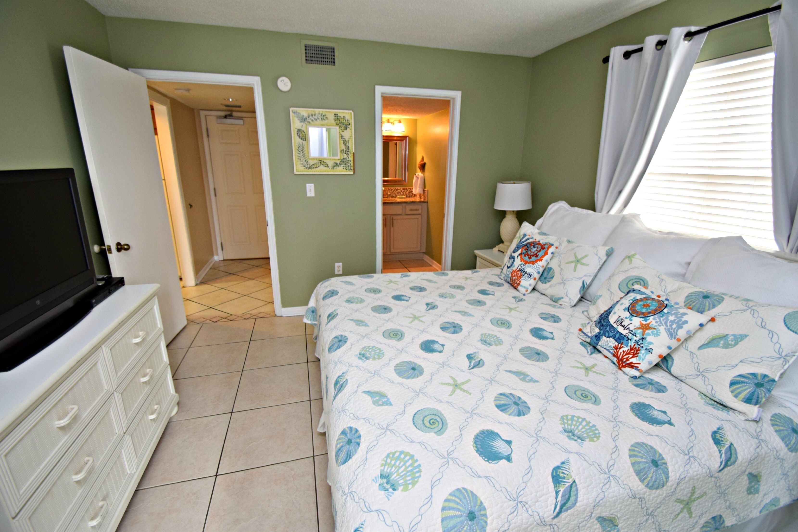 Un dormitorio con cama, cómoda, televisor y baño visible a través de una puerta abierta.