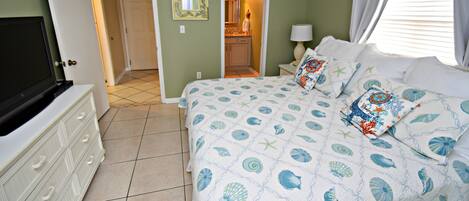 Un dormitorio con cama, cómoda, televisor y baño visible a través de una puerta abierta.