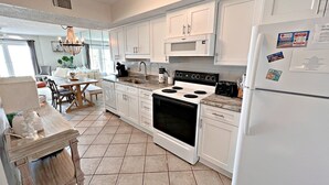 A kitchen with white cabinets, a stove, a refrigerator, and a dining area with a table and chairs. - Surf Side Shores (Gulf Shores)