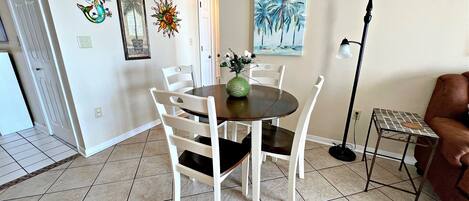 A dining area with a round table, white chairs, and a vase with flowers. There are wall decorations, a painting, and a floor lamp.