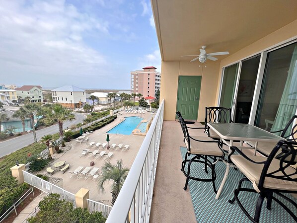 A balcony with a table and chairs, overlooking a swimming pool and a street with palm trees. - The Indies (Gulf Shores)