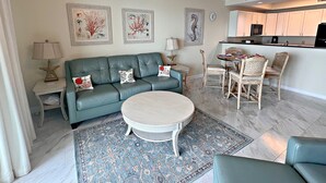 Un salon avec un canapé bleu, une table basse ronde et un tapis à motifs. On y trouve également un coin repas avec une table et des chaises, et une cuisine à l’arrière-plan.