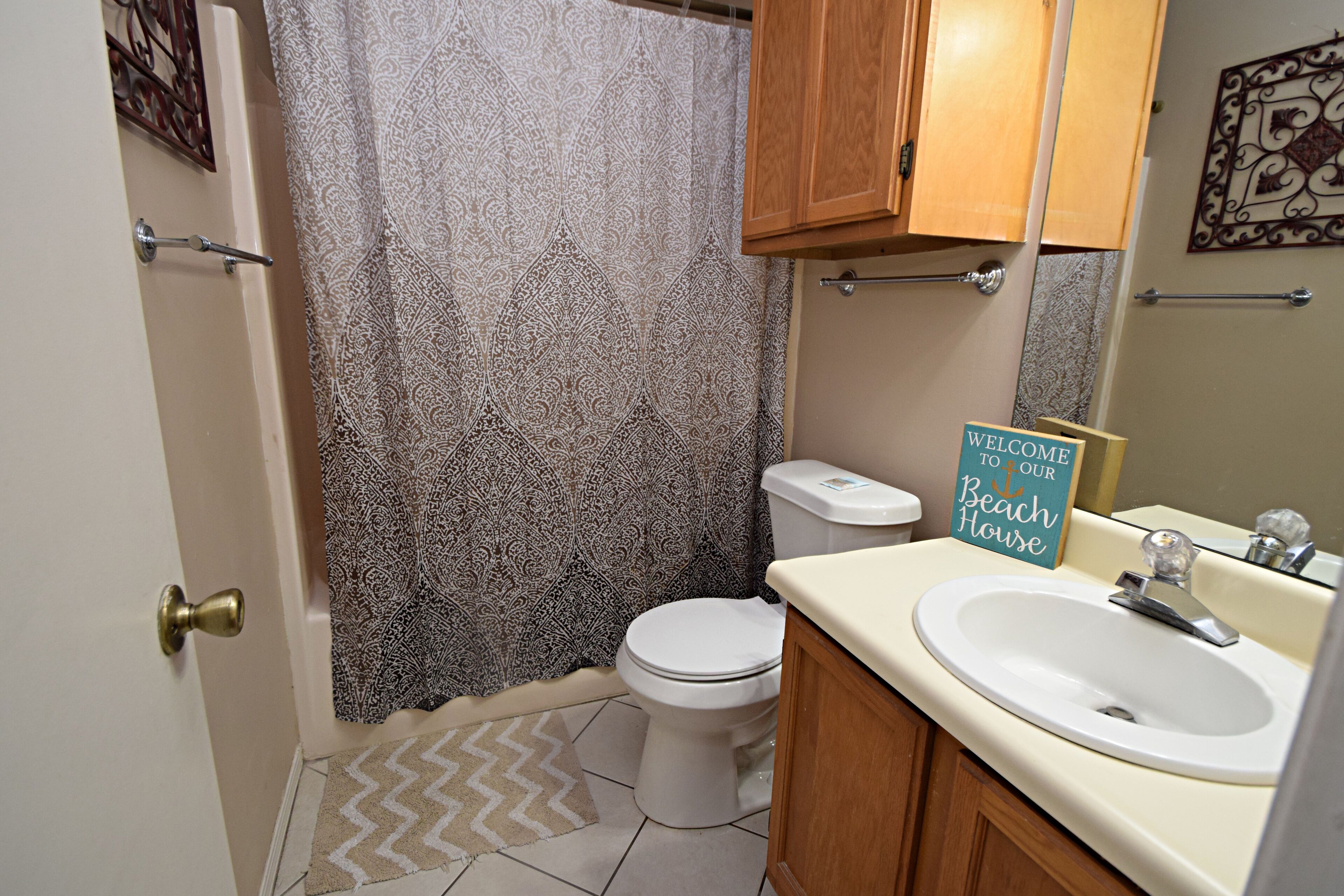 A bathroom with a toilet, a sink, a shower curtain, and a welcome sign.