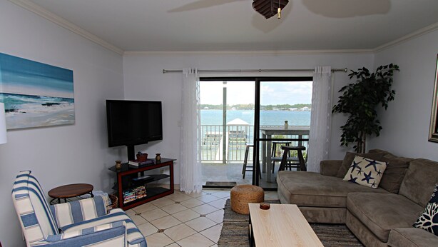 A living room with a sofa, a television, a wooden coffee table, and a view of the sea through a glass door. - The Landing (Gulf Shores)