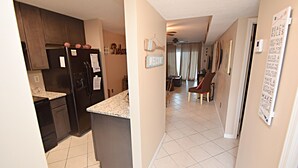 A kitchen with dark cabinets and a granite countertop, a hallway with beige walls and a welcome sign, and a living area with a couch and a coffee table. - The Landing (Gulf Shores)