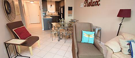A living room with a brown sofa, a glass coffee table, and a dining table with chairs. There is a ceiling fan, a wall clock, and a sign that says "Relax".