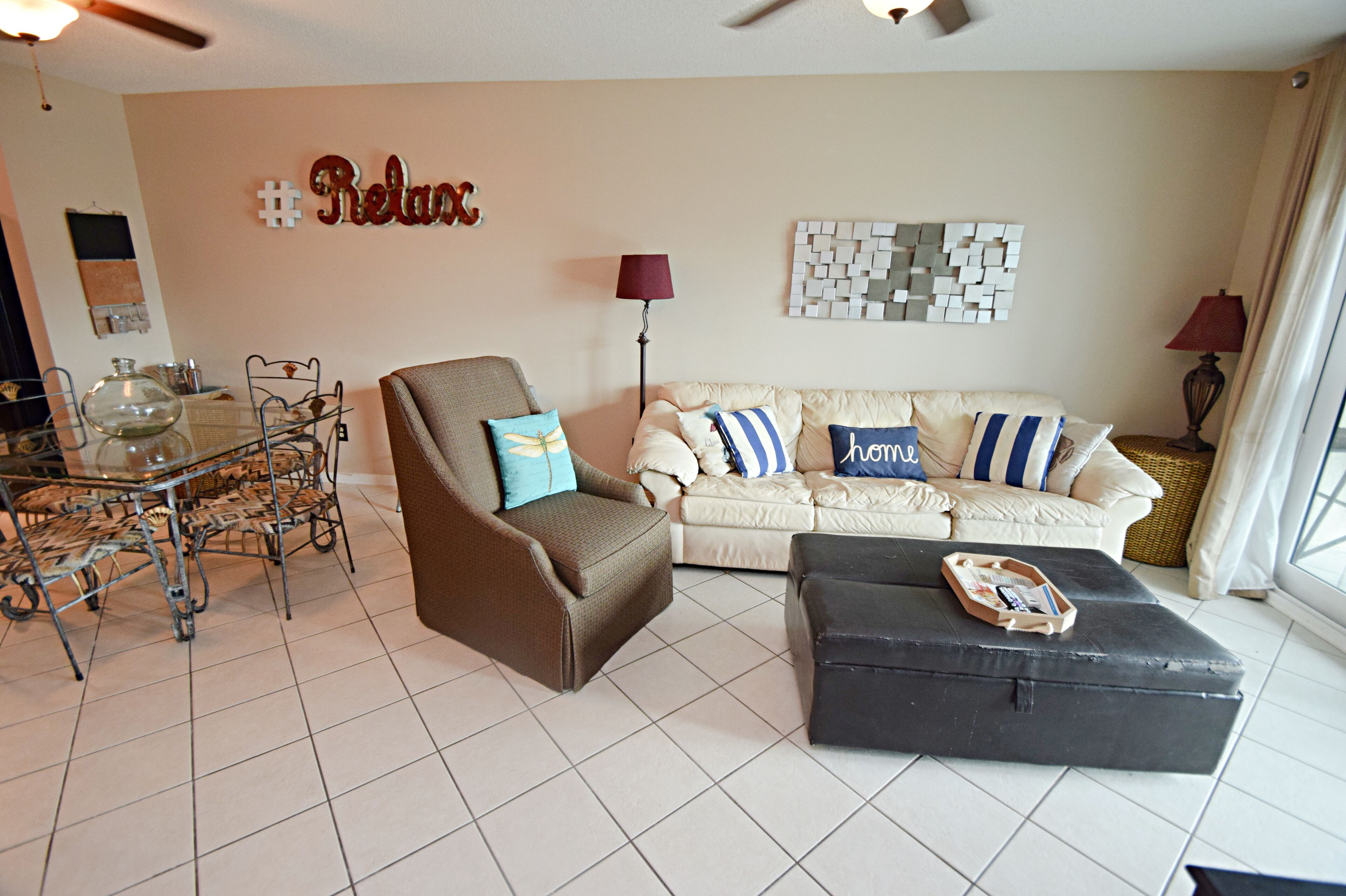 A living room with a brown armchair, a beige sofa with blue and white pillows, a black ottoman, a glass coffee table, and a wall with a '#Relax' sign.