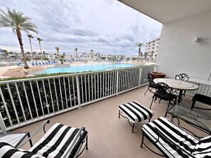 Un balcón con sillones lounge a rayas, una pequeña mesa y vista a un área de piscina con sillas azules y palmeras.