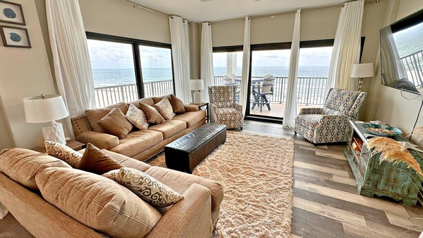 A spacious living room with a large sofa, armchairs, and a coffee table, featuring a view of the ocean through large windows. - The Palms (Orange Beach)