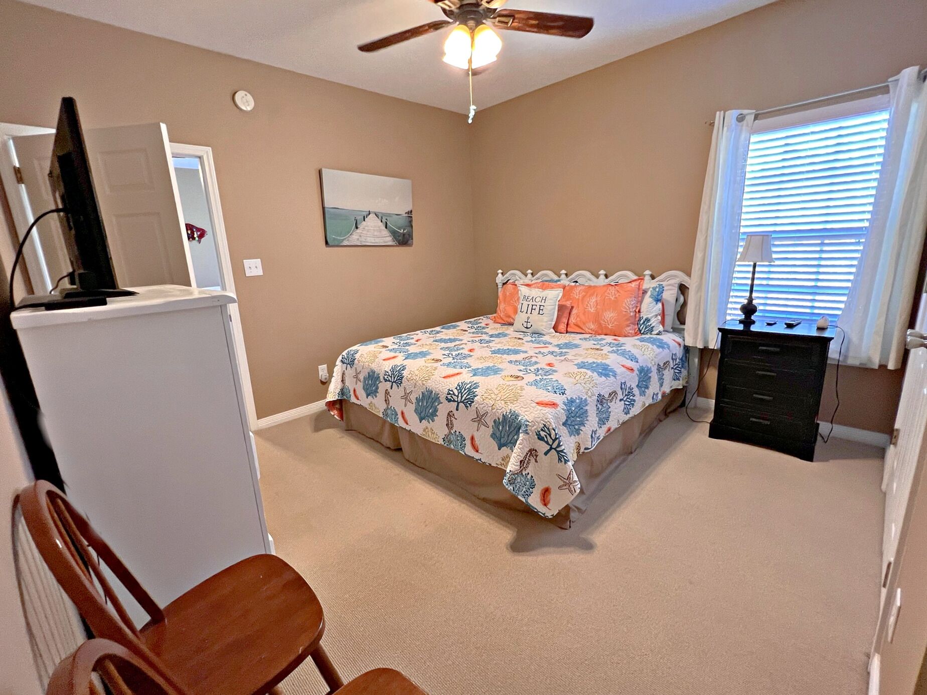 Une chambre à coucher comprenant un lit, un bureau, une chaise, un ventilateur de plafond, une fenêtre avec des stores et un tableau accroché au mur.