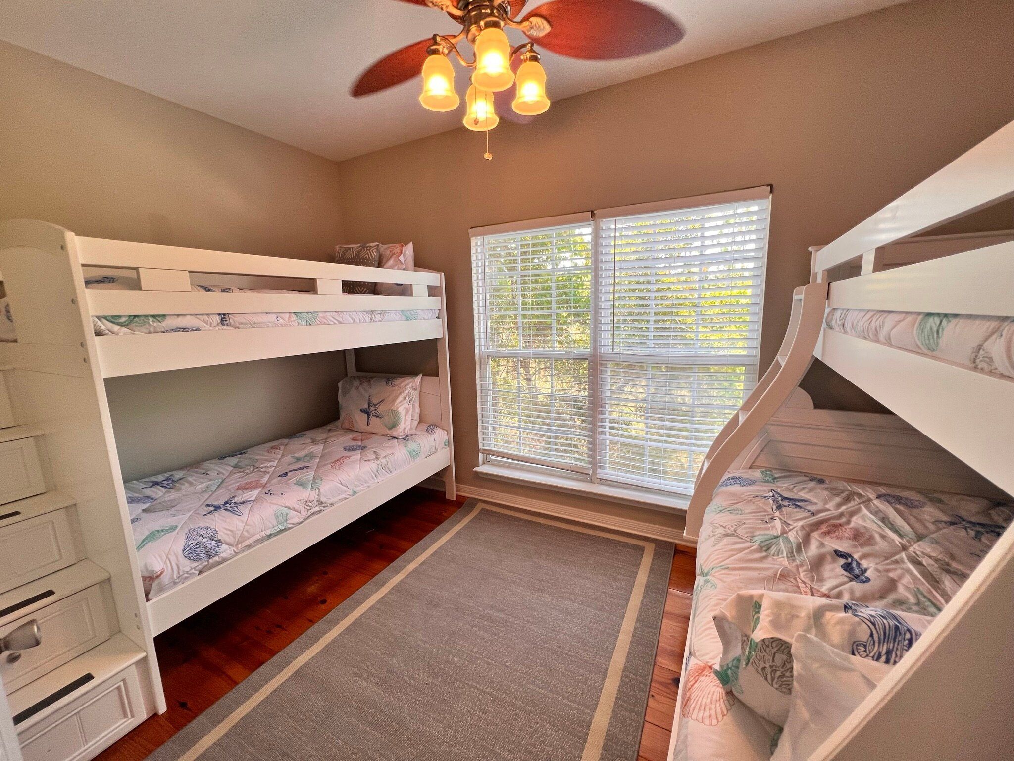 A bunk bed room with a ceiling fan, a window with blinds, and a rug on the floor.