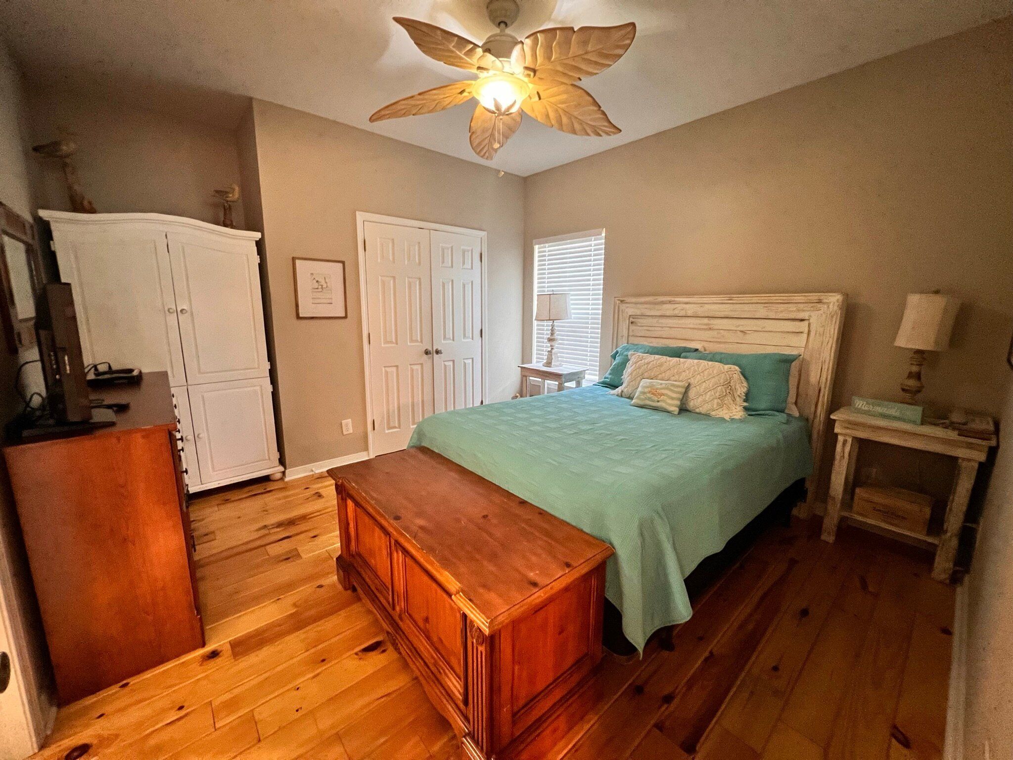 A bedroom with a wooden floor, a bed with a green bedspread, a wooden bench, a white wardrobe, and a ceiling fan.