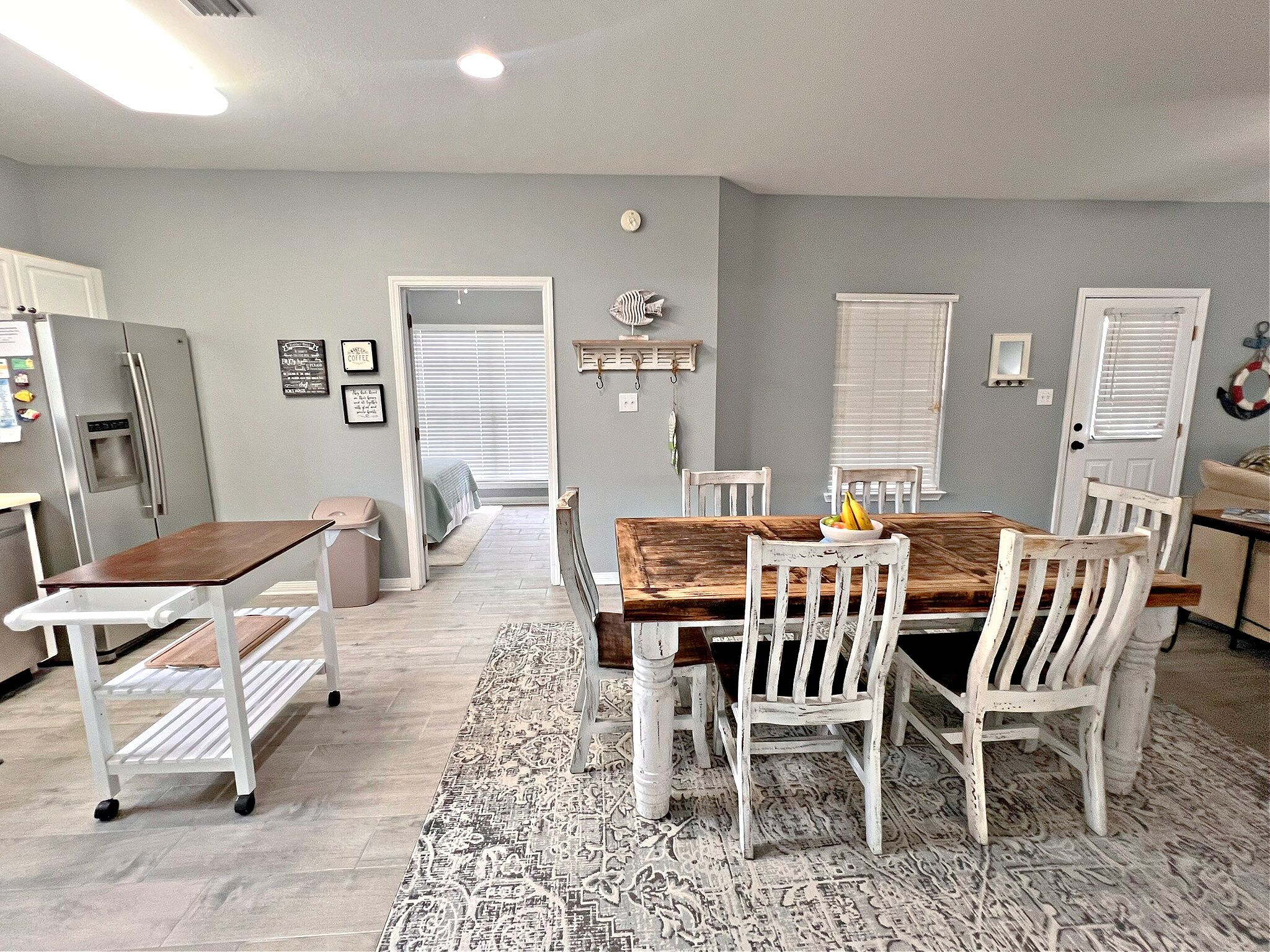 A dining area with a wooden table and white chairs, a kitchen with a refrigerator, and a hallway leading to a bedroom.