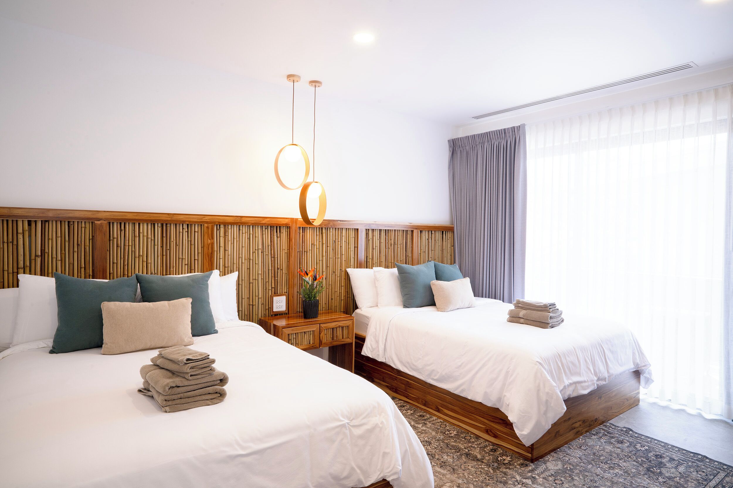 A hotel room with two beds, a wooden headboard, a bedside table with a lamp, and a large window with curtains.