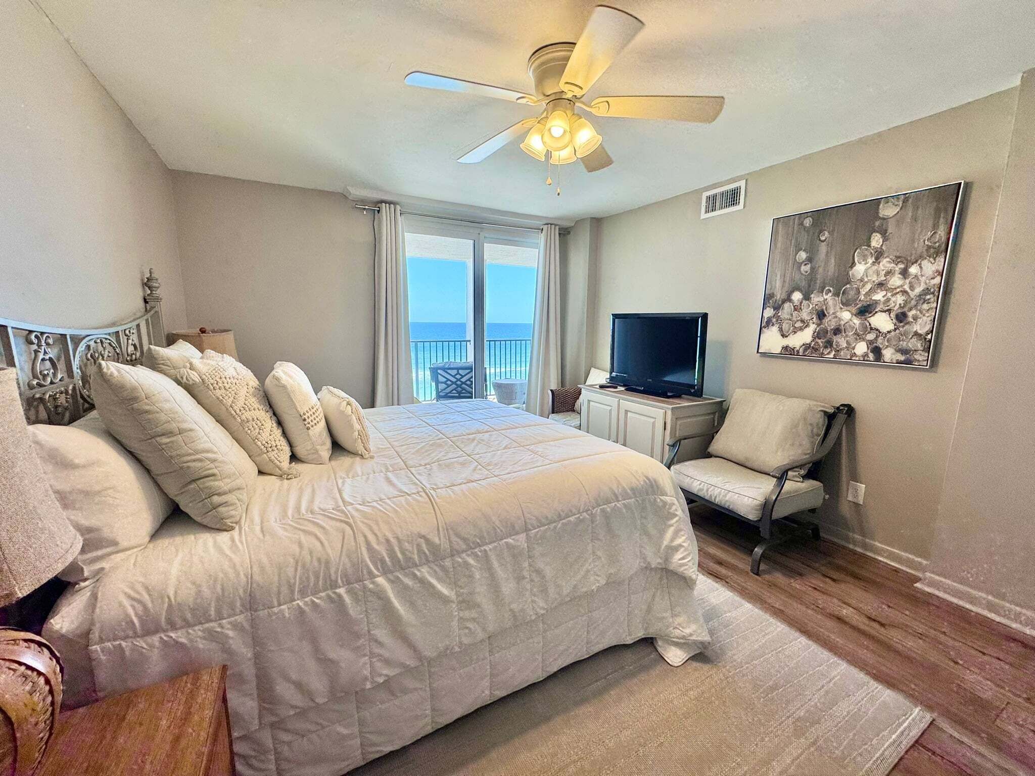 A bedroom with a bed, a ceiling fan, a television, a chair, and a painting on the wall.