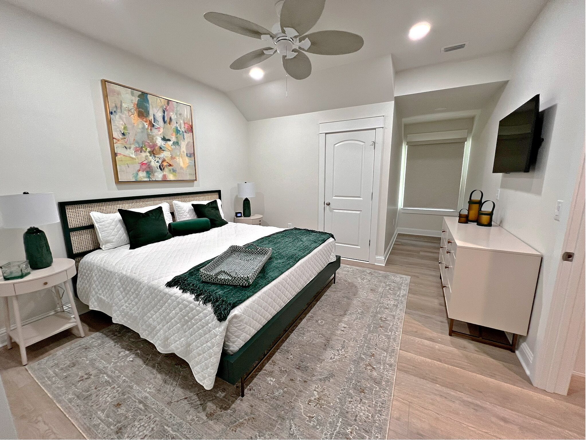 A bedroom with a bed, nightstands, a dresser, a ceiling fan, a painting, and a door.